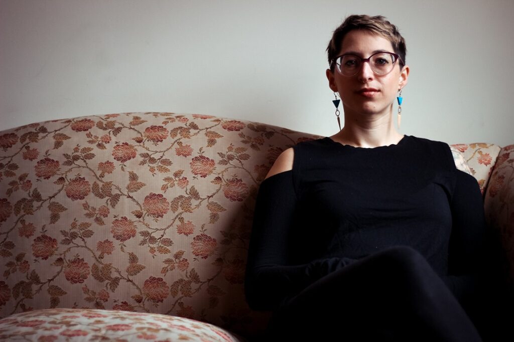 Photo of light-skinned person with short hair and long earrings sitting on a sofa, looking into the camera.