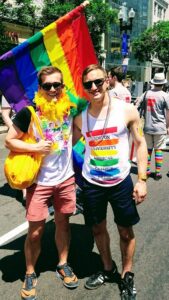 Two young adults side hugging in front of a rainbow flag. They are standing in the street on a sunny day, smiling at the camera and wearing sunglasses.