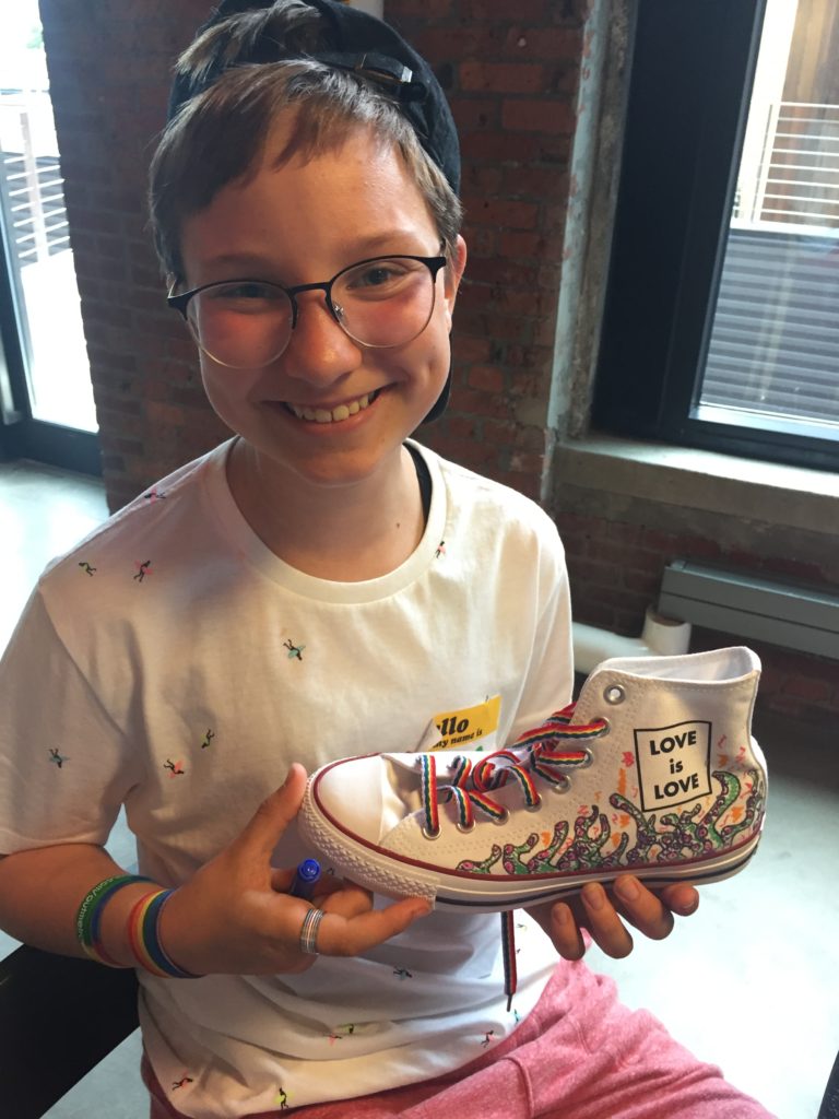 Youth wearing glasses and backward cap, holding Converse high-top that says "Love is love"