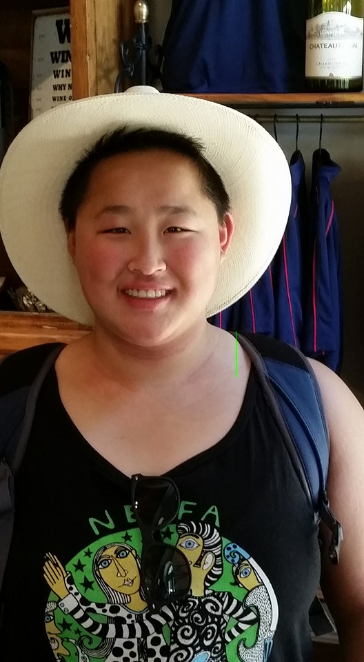 An Asian young adult wearing a large-brimmed hat, black tank top, and backpack smiles into the camera