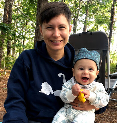 A light-skinned person with short dark hair smiles into camera. She is holding a smiling baby on her lap, and they are sitting in the woods.