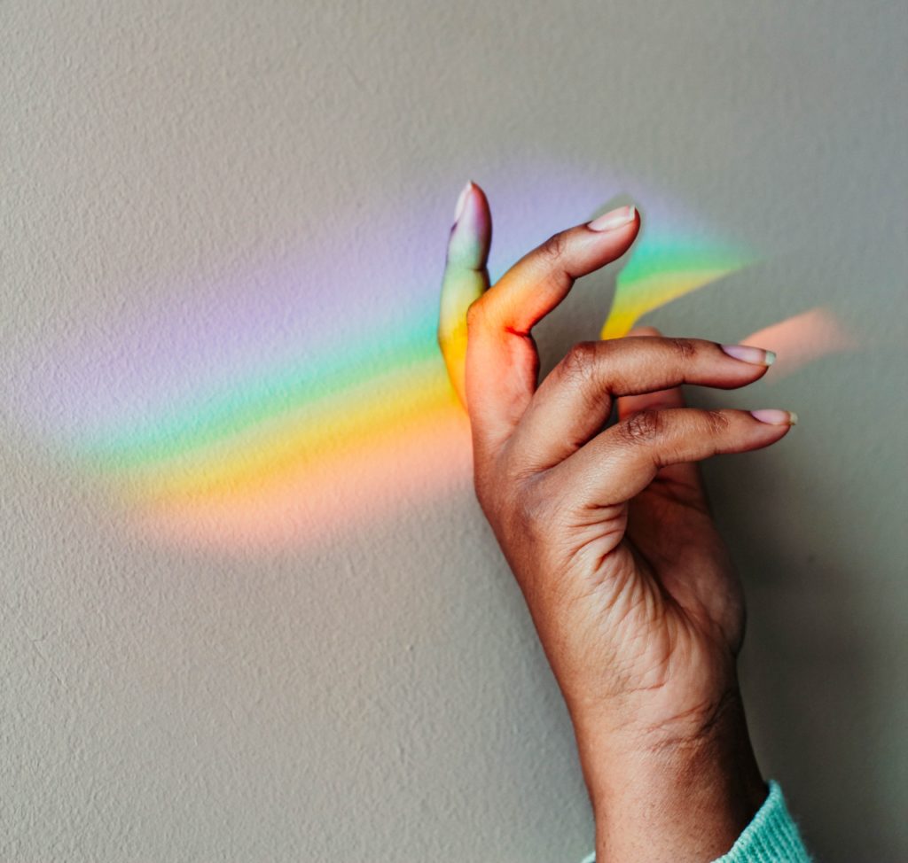 A brown hand gestures in front of a gray wall on which a rainbow has been cast by a prism.