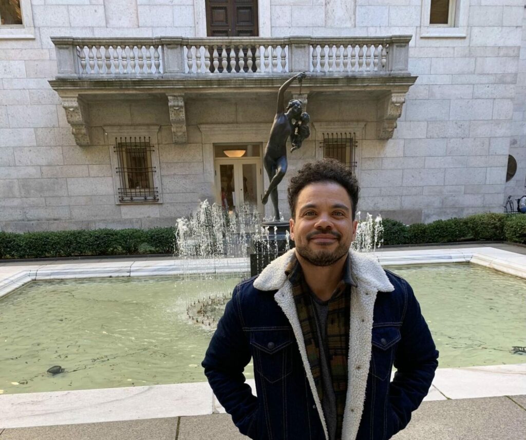 Joel stands in front of a fountain, smiling up into the camera.