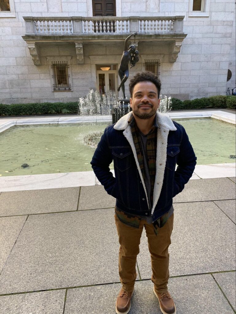 Joel stands in front of a fountain with hands in the pockets of his autumn weight jacket. He has light brown skin and dark, curly, short hair. He is smiling at the camera.